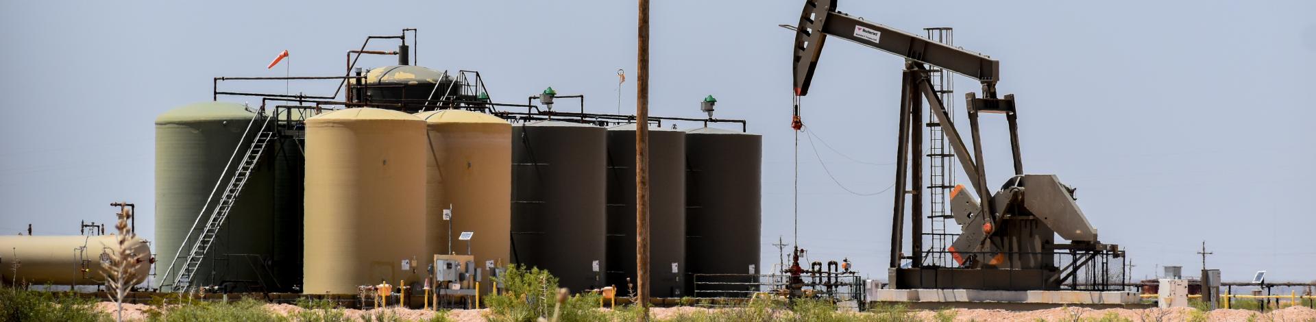 Oil pumpjack operating next to large storage tanks in a dry, desert landscape under a clear sky.